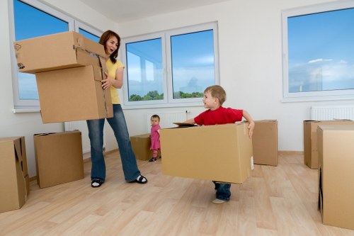 A woman in a yellow top and jeans is inside a bright, spacious room with large windows showing blue sky and green trees outside, carrying a light brown cardboard box. Two children are present: a young girl in a pink outfit standing near the woman, and a young boy in a red shirt and jeans, holding and lifting a medium-sized cardboard box. Several other boxes of various sizes, all made of brown cardboard, are placed around the room, some stacked and some on the floor. The room has light wooden flooring and white walls, indicating a residential space, possibly in the process of moving or packing, with natural daylight streaming through the windows. The scene depicts a household moving activity, with the woman and children engaged in handling belongings before relocation in Hounslow, demonstrating typical house removal procedures with household boxes and minimal furnishings present.