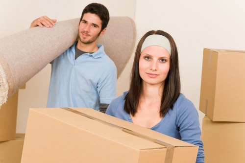 A young man and woman, both dressed in casual clothing, are inside a residential property preparing for a house move. The man, wearing a light blue polo shirt, is standing behind the woman and lifting a large, light grey upholstered sofa with a textured fabric surface. The woman, with long dark hair and wearing a white headband, is facing forward and holding a medium-sized, brown cardboard box with visible flaps, suggesting it is sealed for moving. Behind them, there are additional cardboard boxes stacked on a beige or neutral-walled interior, indicating an organized packing process. The scene takes place in what appears to be a living room or hallway with soft, natural light illuminating the space, highlighting the textures of the furniture and boxes. The background features a clear view of the room's open layout, emphasizing the preparatory stage of a house relocation, consistent with professional house removals services such as those offered by Hounslow Man with Van in the Hounslow area, close to postcode TW or TW3. The focus remains on the careful handling of household belongings, essential for safe and efficient removals in the local area.