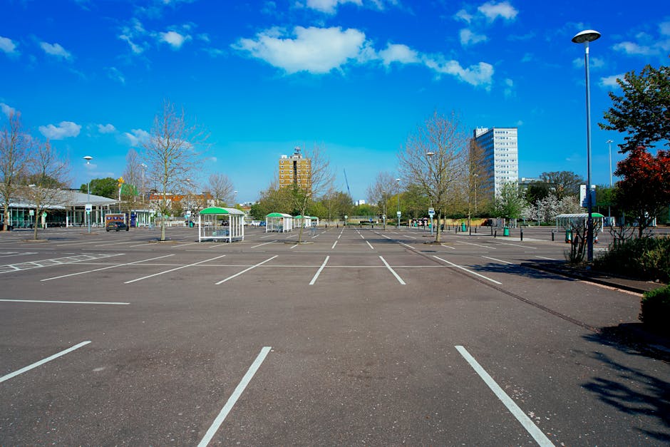 A wide view of an empty outdoor parking lot with clearly marked white lines for parking spaces, set against a backdrop of leafless and blossoming trees and several multi-storey office buildings under