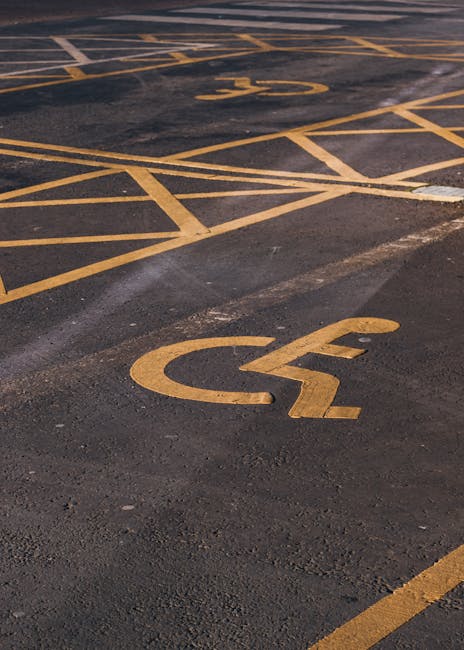 A wide view of an empty outdoor parking lot with clearly marked white lines for parking spaces, set against a backdrop of leafless and blossoming trees and several multi-storey office buildings under