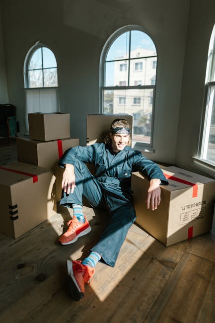 A man with a beard and wearing a black cap and white polo shirt is holding a cardboard box during a home relocation process. He is standing outside a moving van with its rear door open, loaded with mu